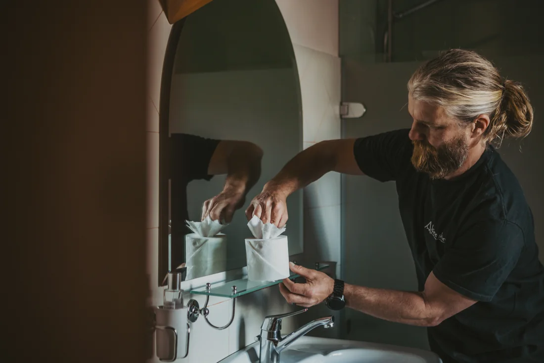 Dan folding a toilet-paper flower as a finishing touch in a chalet bathroom